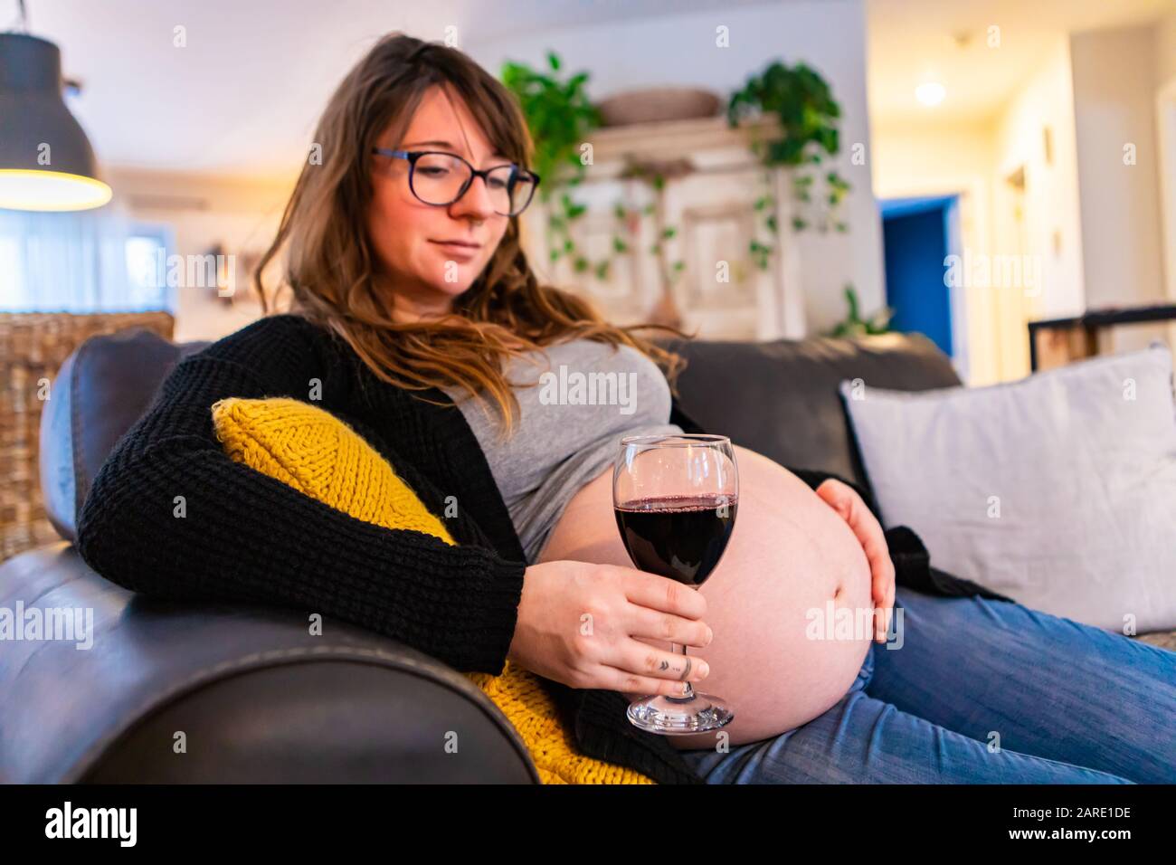A portrait of a heavily pregnant woman sitting on a sofa in the home with a glass of red wine, unaware of the risks of fetal alcohol syndrome. Copy space to right Stock Photo