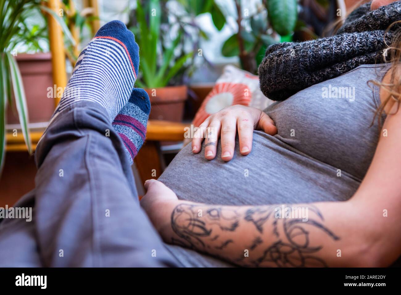 A closeup shot on the feet of a man resting over heavily pregnant partner with large swollen stomach and tattooed arms. Parents await arrival of child Stock Photo