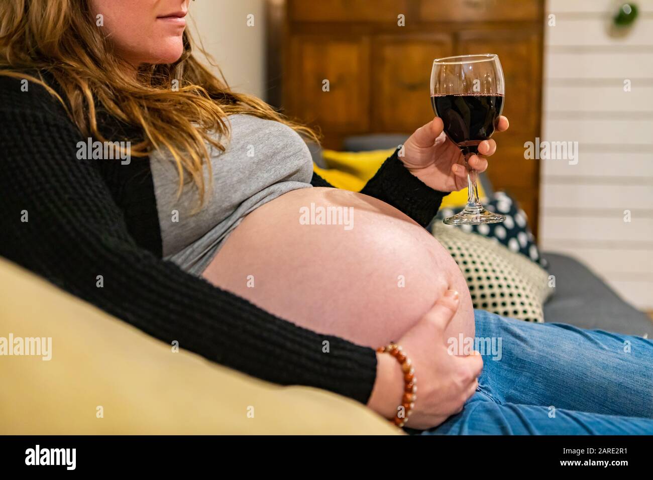 A heavily pregnant woman nearing labor is seen sitting on a sofa holding swollen tummy with one hand and a glass of red wine in other, with copy space Stock Photo