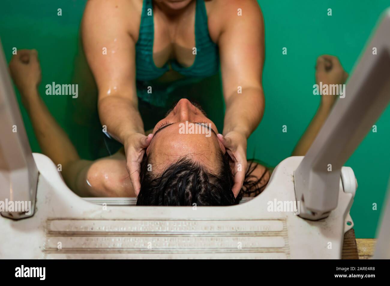 High angle close-up view of female therapist hand giving water massage on head to young man. leaning on swimming pool ladder during water therapy Stock Photo