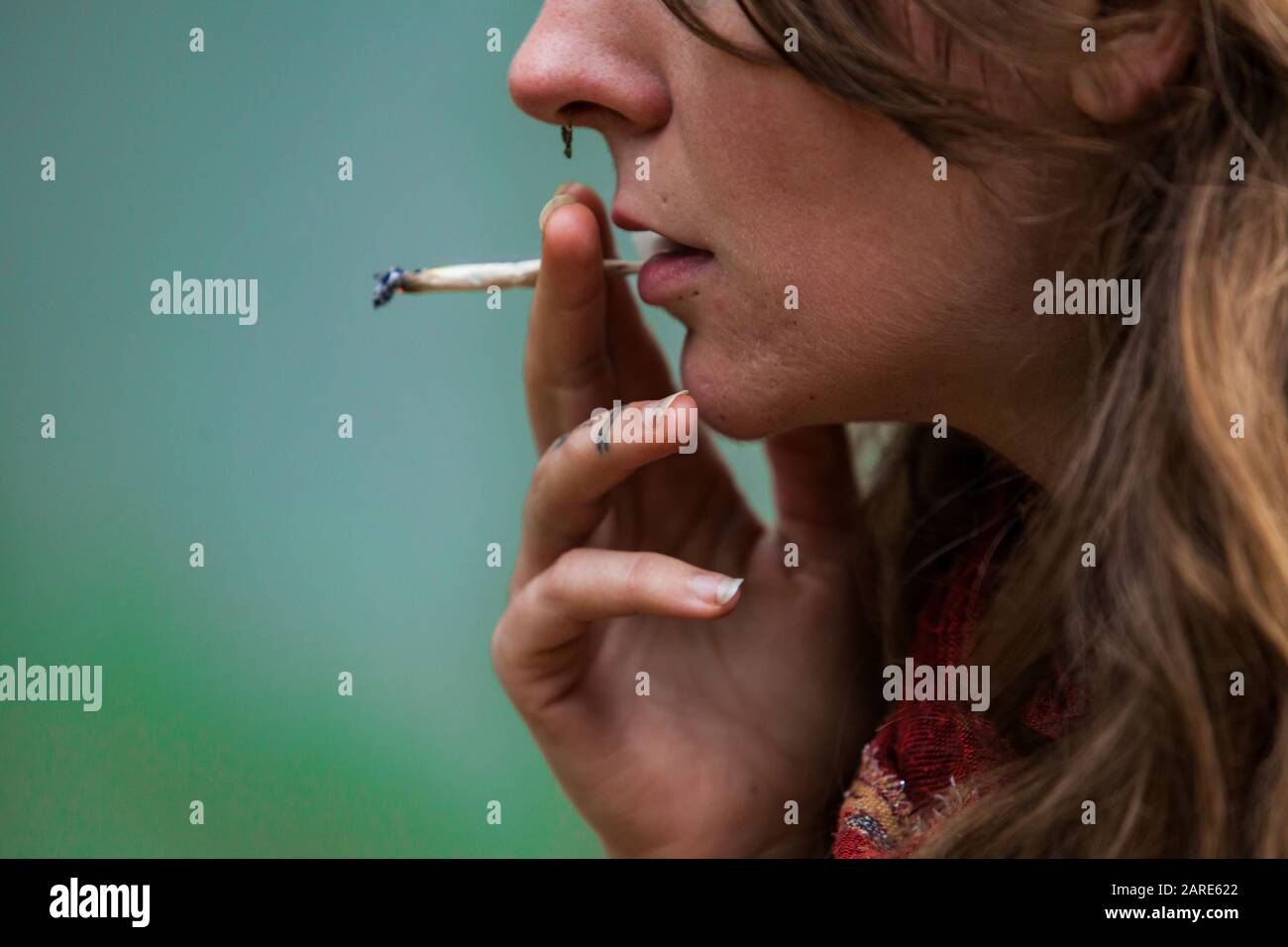 Closeup cropped shot of lower half of caucasian woman's face smoking hand rolled marijuana joint. Cannabis cigarette held by woman's tattooed fingers  Stock Photo