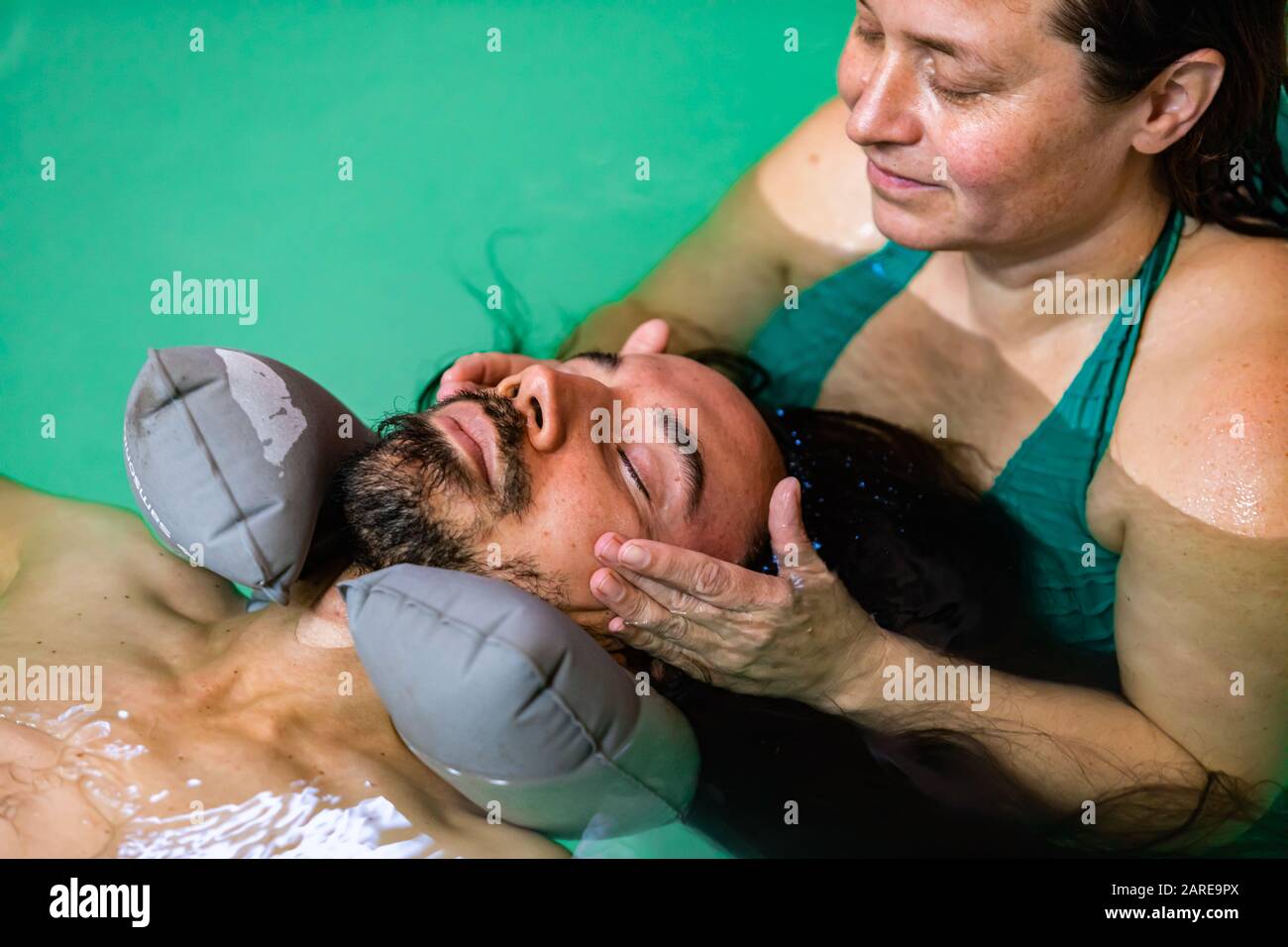 Close-up of young man with closed eyes wearing rubber neck pillow. around his neck getting spa water massage treatment from trainer in swimming pool Stock Photo