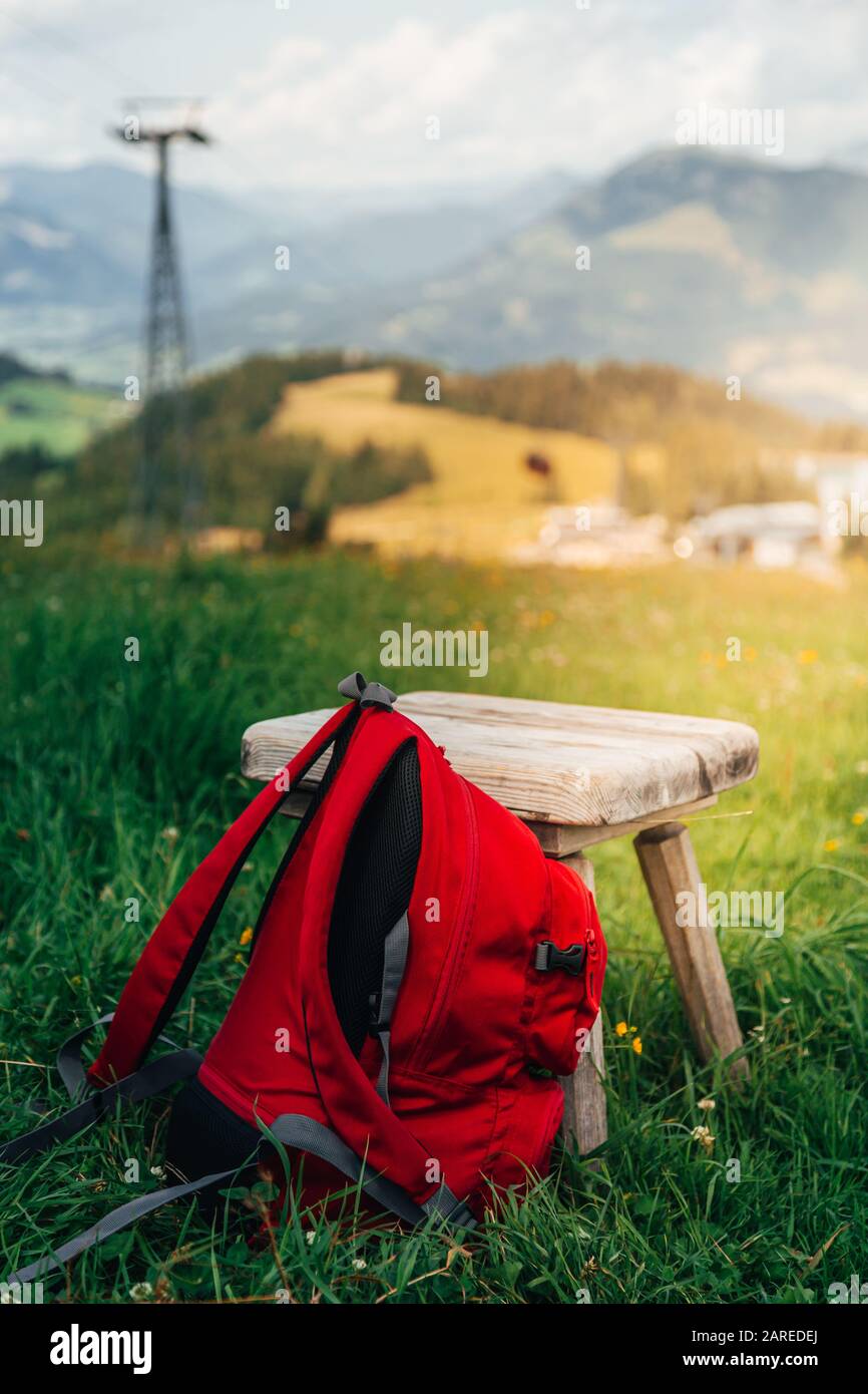 Red backpack lying against a wooden stool in an alpine pasture in the alps with mountains in the background. Stock Photo