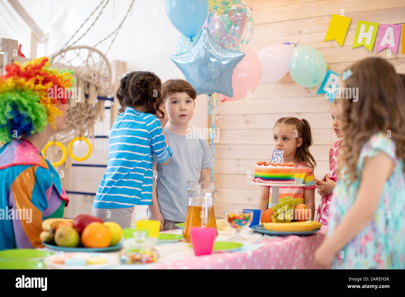Group of adorable kids standing around festive table at party. Little girl congratulating birthday boy. Stock Photo