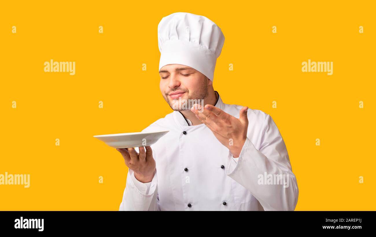 Professional Chef Holding Empty Plate Smelling Invisible Dish, Yellow Background Stock Photo