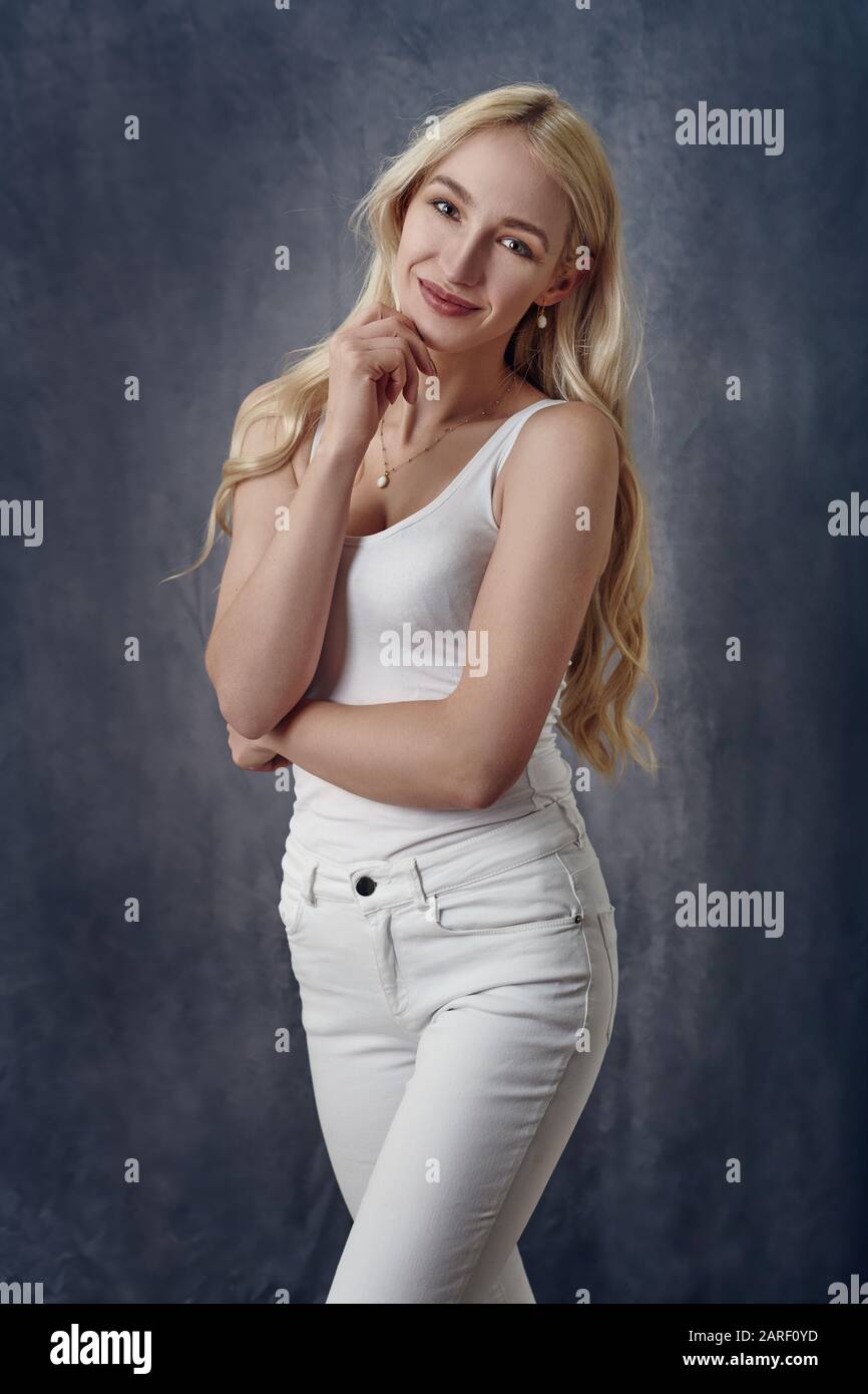 Attractive sexy slender blond woman with long hair posing in off white summer top and jeans with her hand to her chin smiling at the camera over a gre Stock Photo