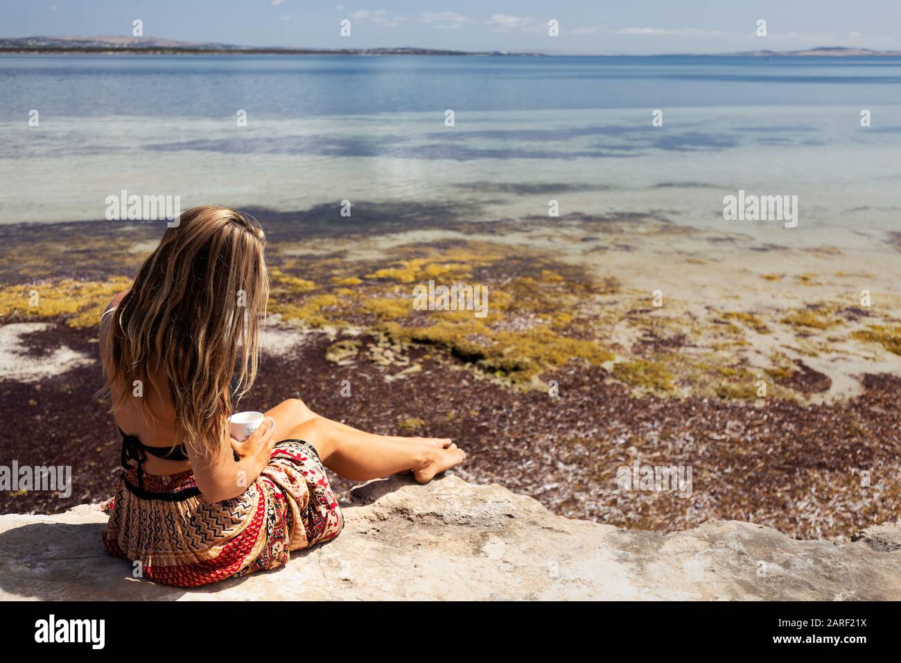 Beautiful bohemian woman relaxing while drinking cup of tea by the sea on bright, sunny day in South Australia. Stock Photo