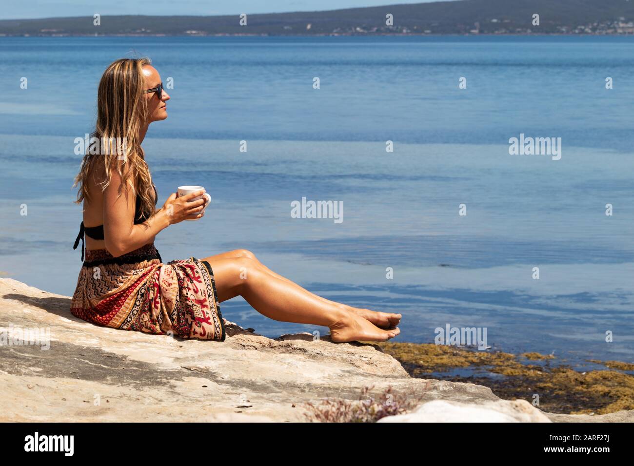 Beautiful woman relaxing while drinking cup of tea by the sea on sunny, summer day in South Australia. Stock Photo
