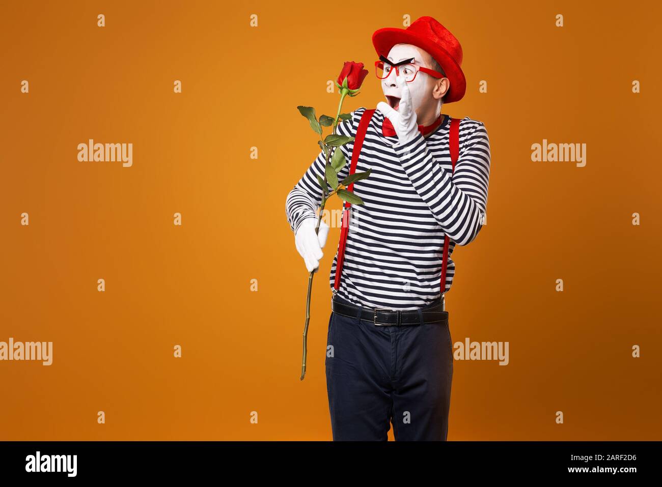 Sad mime with white face in red hat and striped t-shirt holding rose on empty orange background in studio Stock Photo