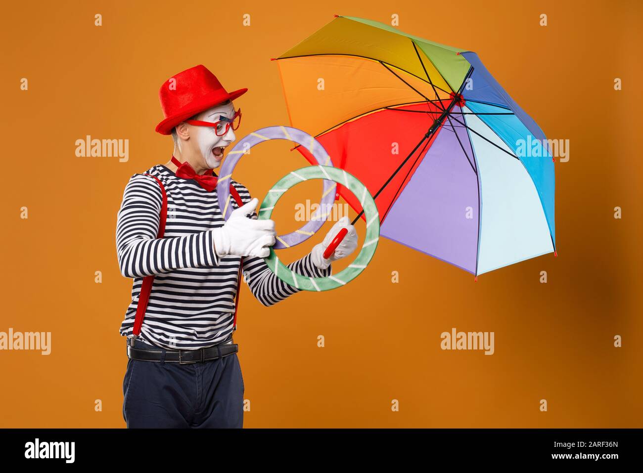 Cheerful mime with rings and multi-colored umbrella on empty orange background in studio Stock Photo