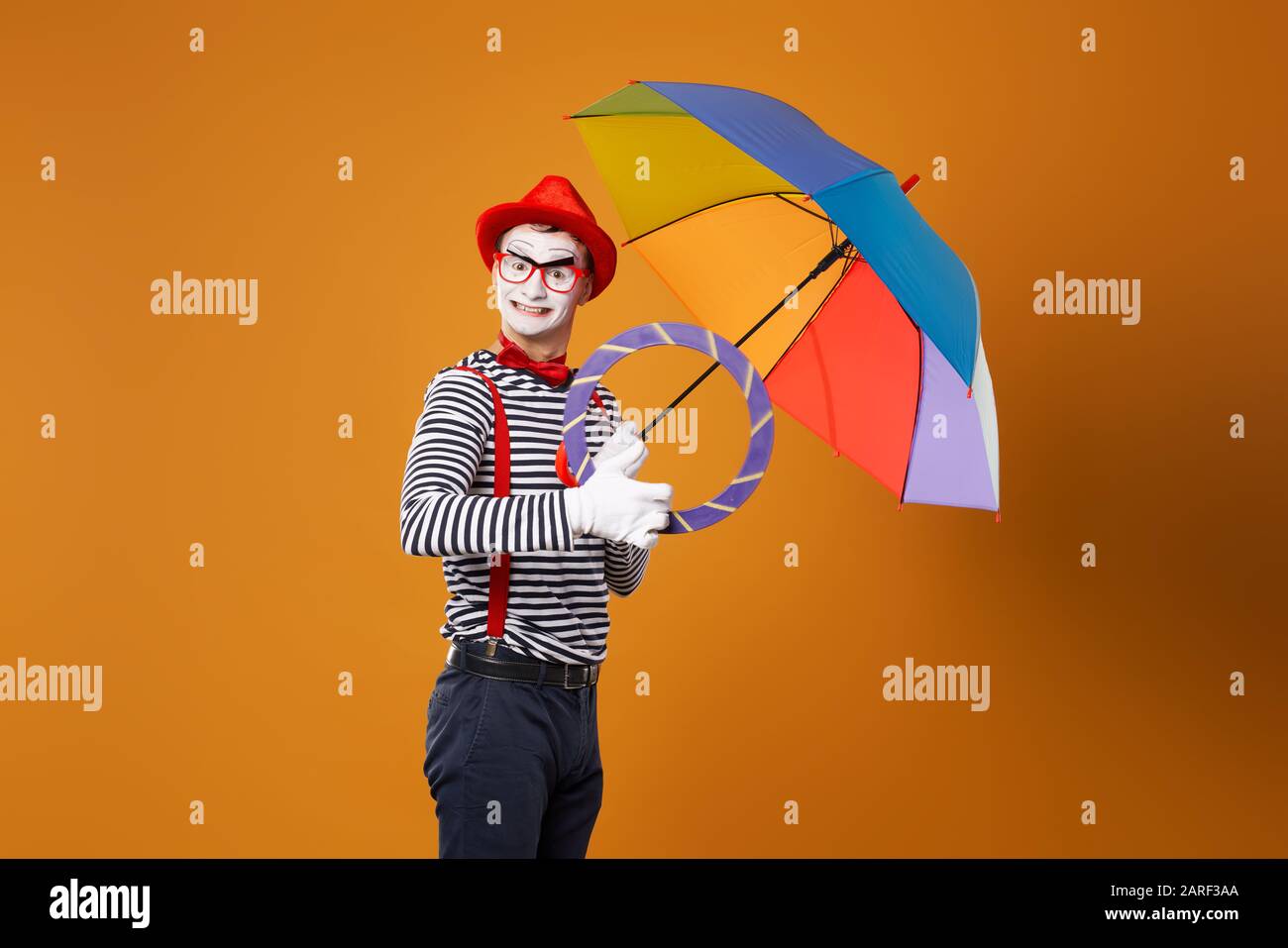 Funny mime with multi-colored umbrella on empty orange background in studio Stock Photo