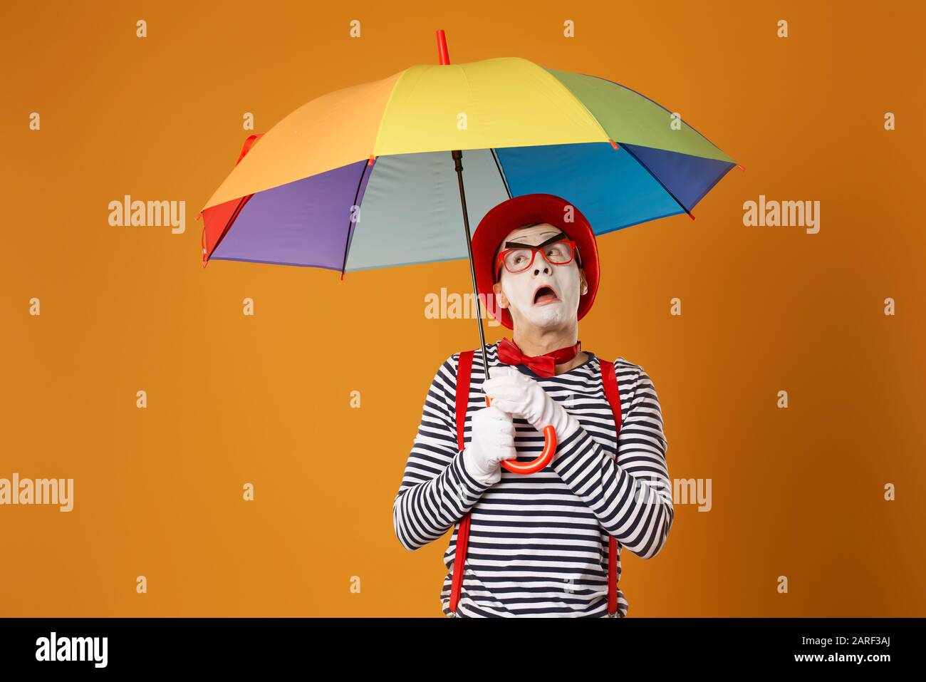 Surprised mime with multi-colored umbrella on empty orange background in studio Stock Photo
