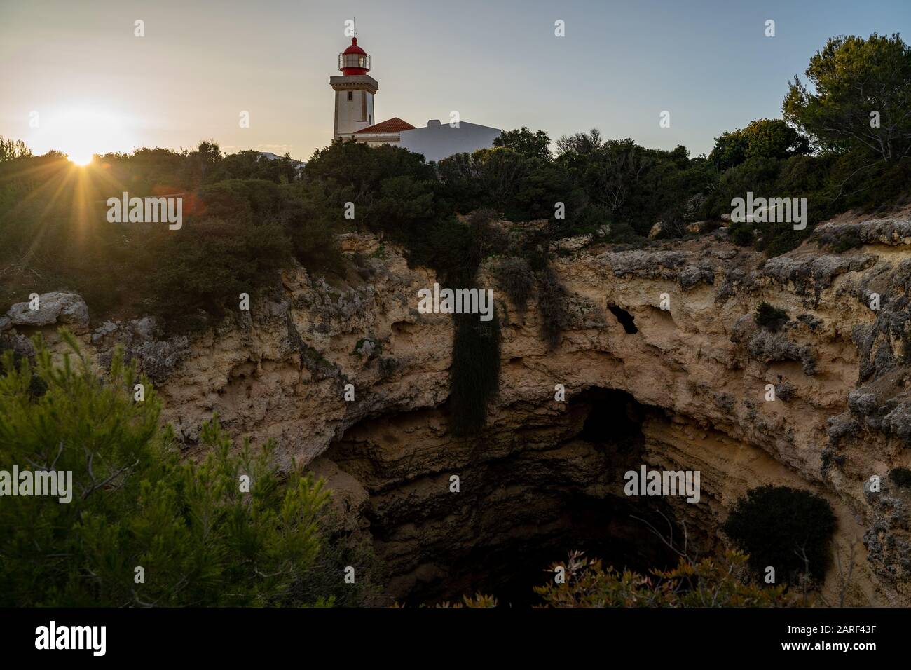 Alfanzina Lighthouse, as seen framed by trees, sunflare and sea caves at sunset in the Algarve region of Portugal Stock Photo