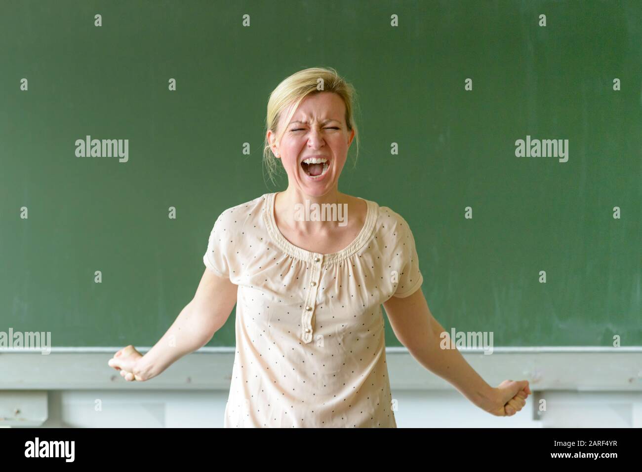 Emotional angry female school teacher yelling at her class while clenching her fists in a show of temper in front of a chalkboard Stock Photo