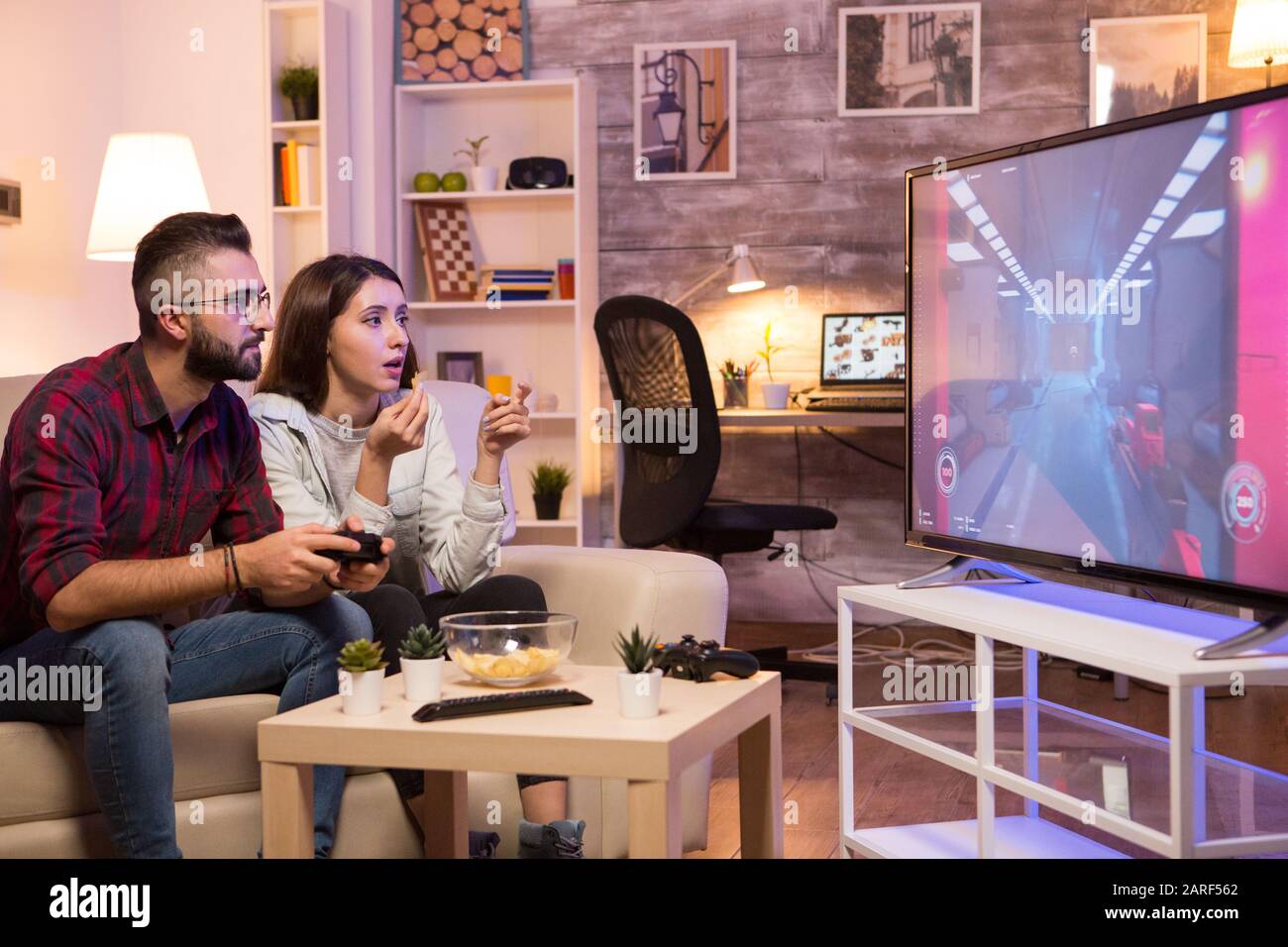Boyfriend playing video games on television using controller while girlfriend is eating chips. Couple sitting on sofa. Stock Photo