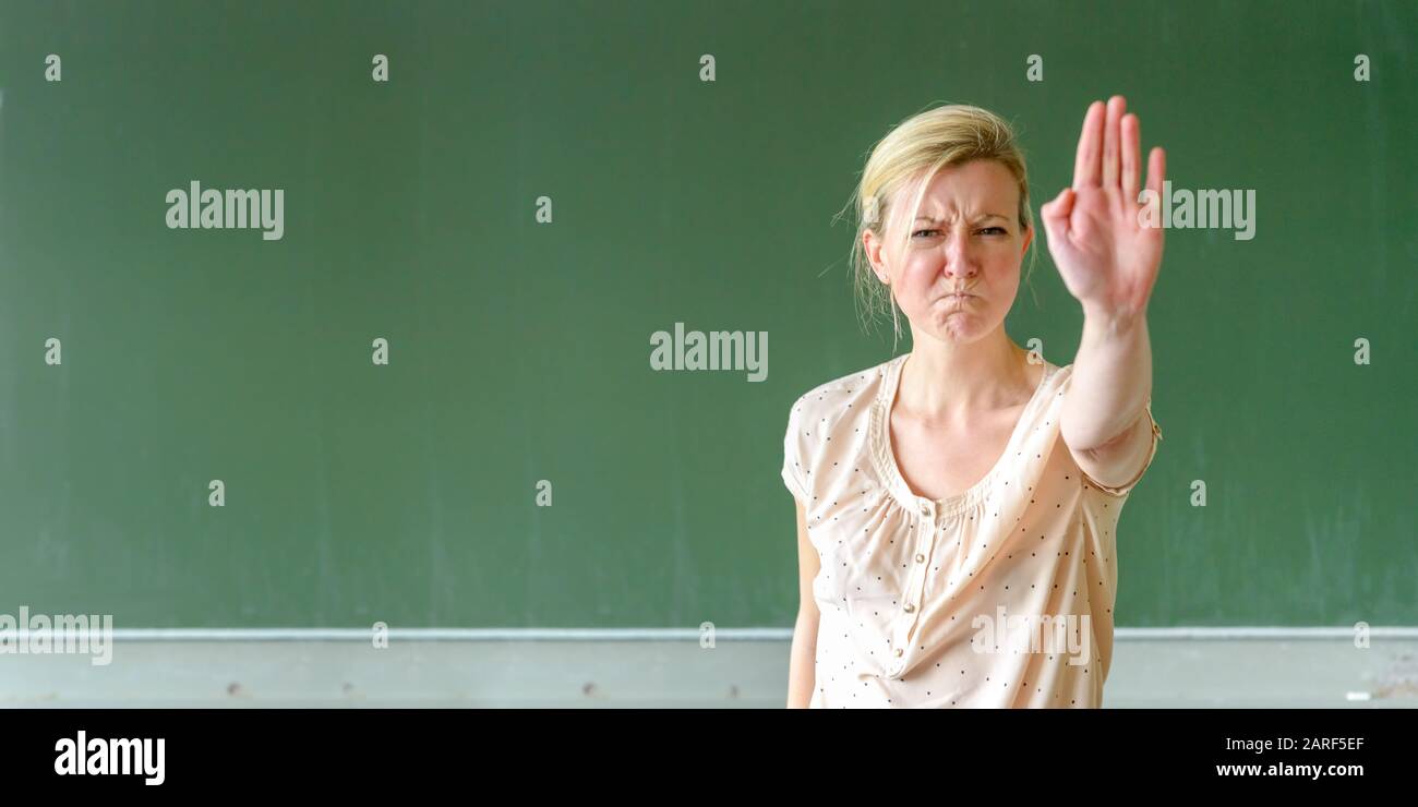Angry teacher or lecturer calling a halt by gesturing with her hand while screwing up her face in frustration in front of a chalkboard Stock Photo