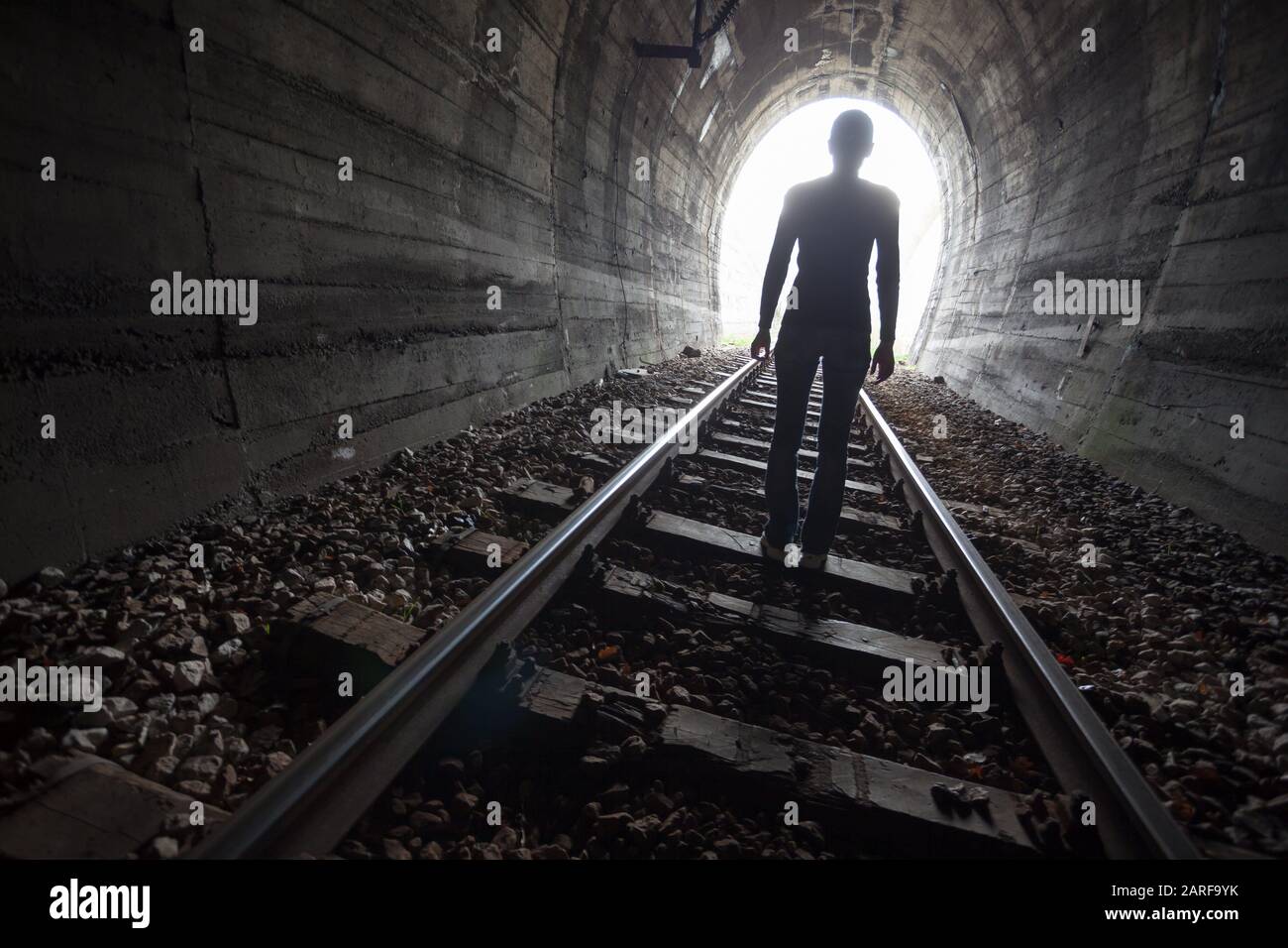 Man silhouetted in a tunnel standing in the center of the railway tracks looking towards the light at the end of the tunnel in a conceptual image. Stock Photo