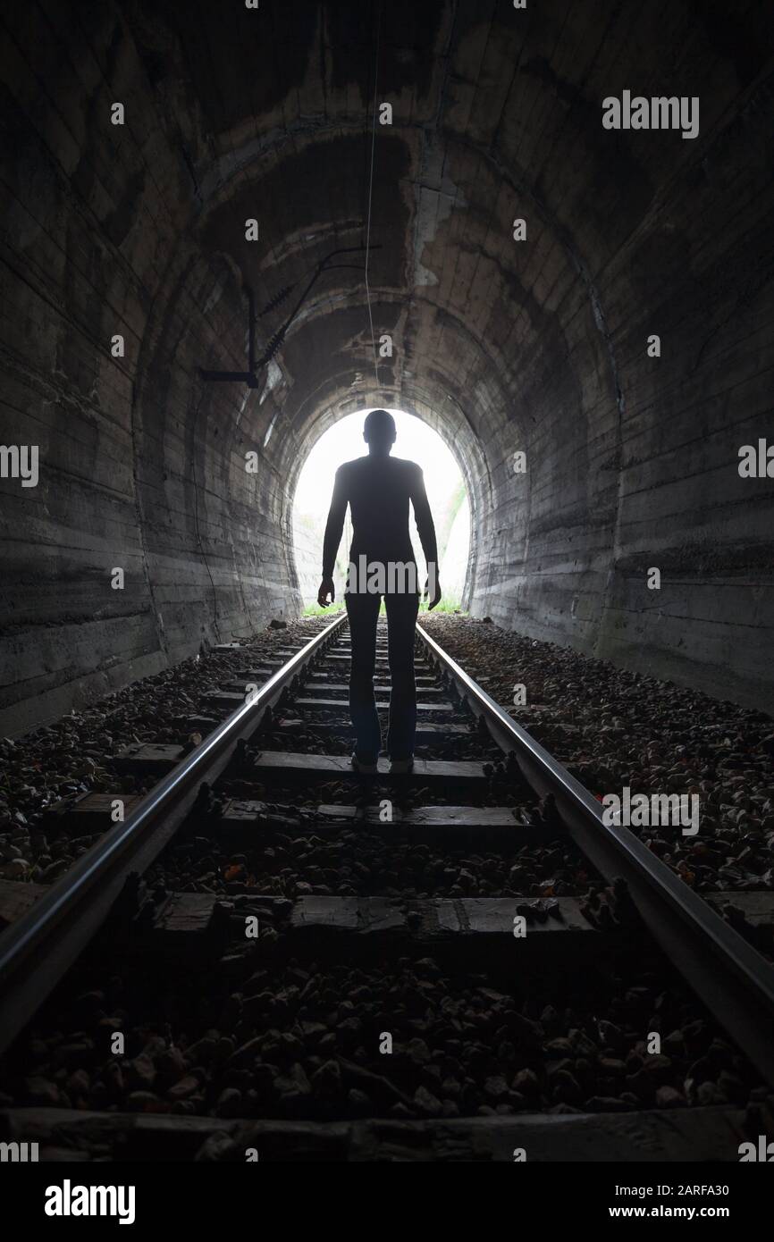 Man silhouetted in a tunnel standing in the center of the railway tracks looking towards the light at the end of the tunnel in a conceptual image. Stock Photo