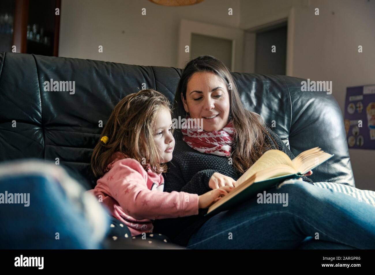 Single parent family concept. Mother and daughter reading a book on the sofa in the living room. Empty copy space for Editor's text. Stock Photo