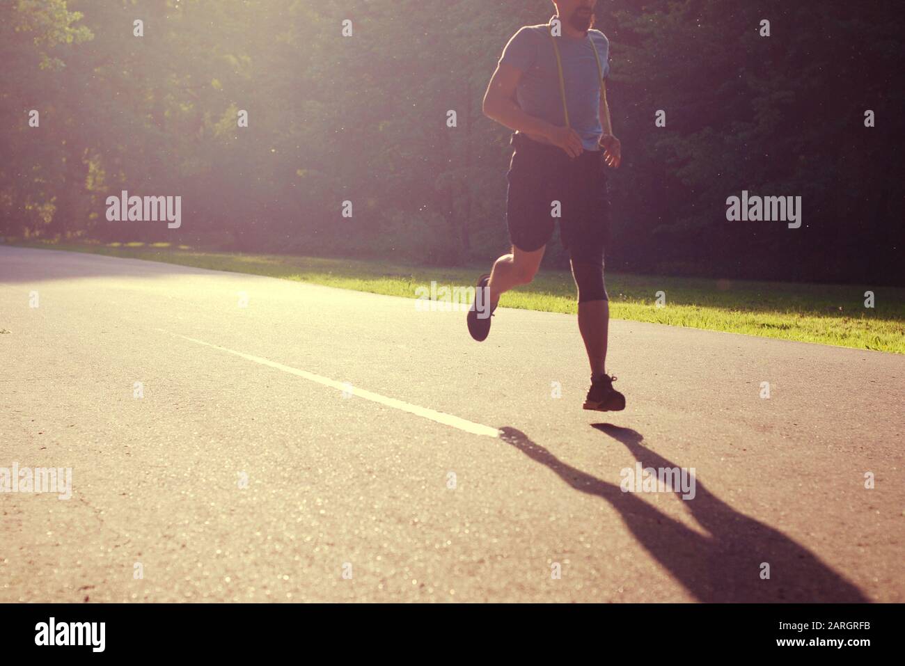 Man jogging in summer park, silhouette of running guy in backlight casting shadow on the asphalt path. Stock Photo