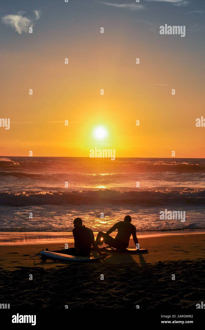 Silhouettes of two surfers sitting on the Beach at Sunset Stock Photo ...