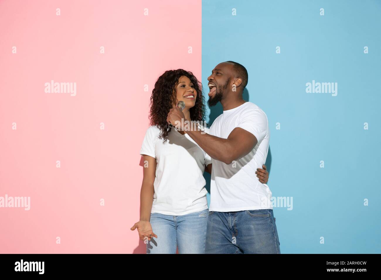 Singing song together. Young man and woman in casual clothes on pink, blue bicolored background. Concept of human emotions, facial expession, relations, ad. Beautiful african-american couple. Stock Photo