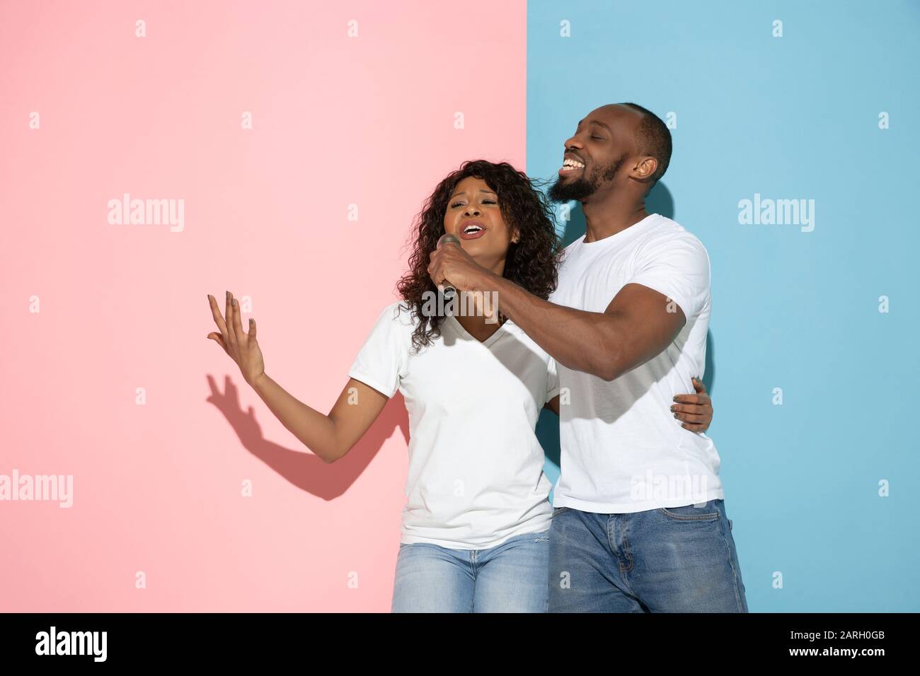 Singing song together. Young man and woman in casual clothes on pink, blue bicolored background. Concept of human emotions, facial expession, relations, ad. Beautiful african-american couple. Stock Photo