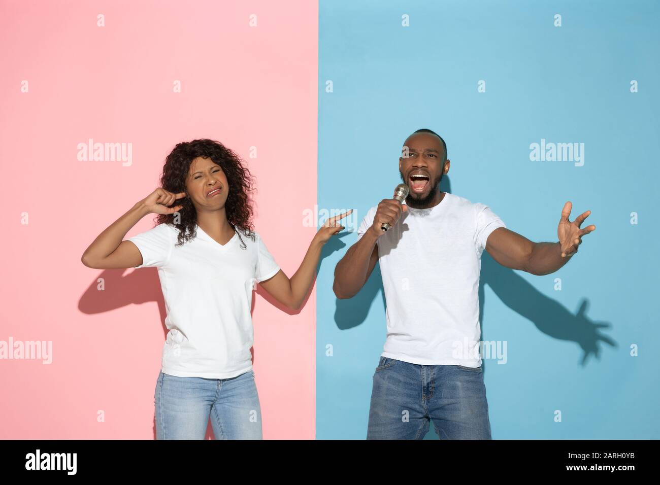 He singing, she angry. Young man and woman in casual clothes on pink, blue bicolored background. Concept of human emotions, facial expession, relations, ad. Beautiful african-american couple. Stock Photo