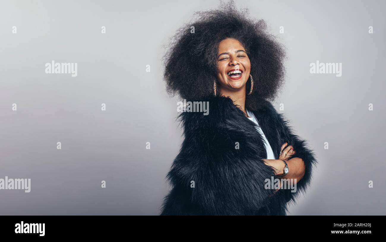 Smiling woman in afro hairstyle standing against grey background. Portrait of cheerful woman in fur coat looking at camera. Stock Photo