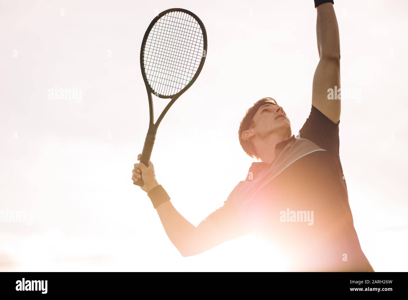 Young tennis player about to hit the ball on a sunny day. Professional tennis player making a serve with bright sunlight from behind. Stock Photo