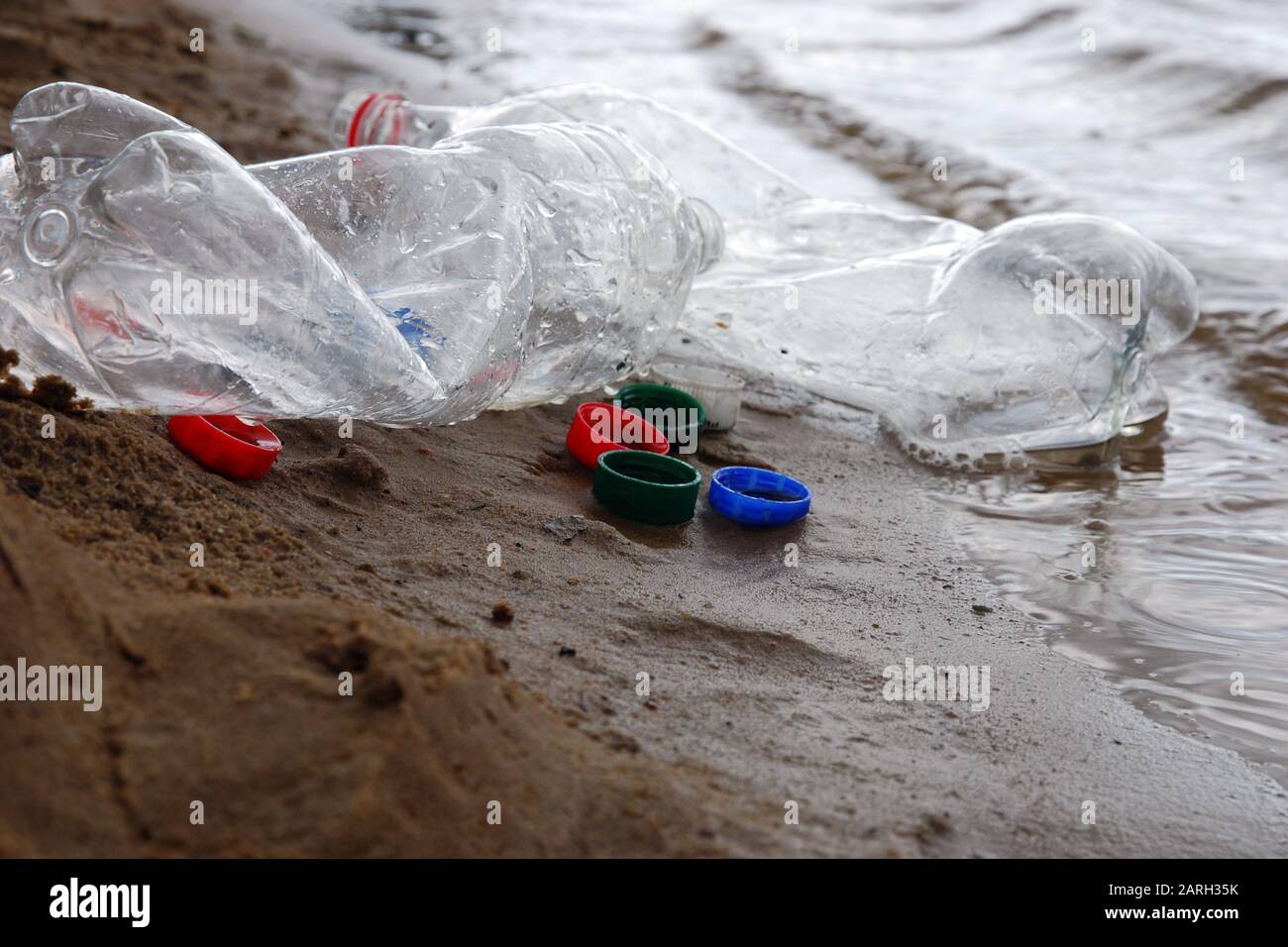 Plastic waste left by campers at river or lake shore, bottles and plastic bottle caps on the sand in the water close up Stock Photo
