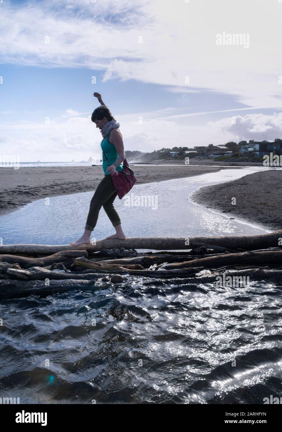 A woman crosses a stream by balancing on a large driftwood log, at Oakura Beach, New Zealand Stock Photo