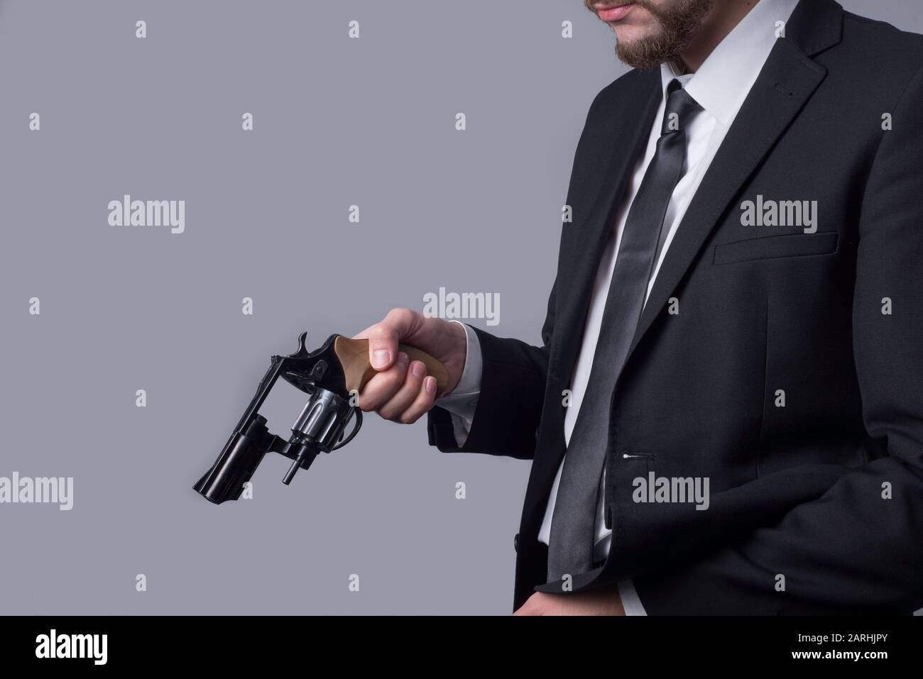 Portrait of a bearded man in a business suit holds a revolver in his hand, reloading it. On a gray background. Criminal type man, gangster, killer, bu Stock Photo