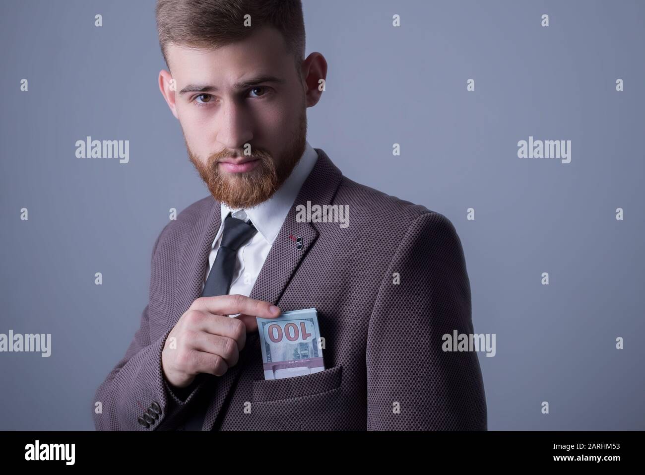 Studio portrait of a young bearded handsome guy of twenty-five years old, in an official suit, looking at the camera, holding a press with dollar bill Stock Photo