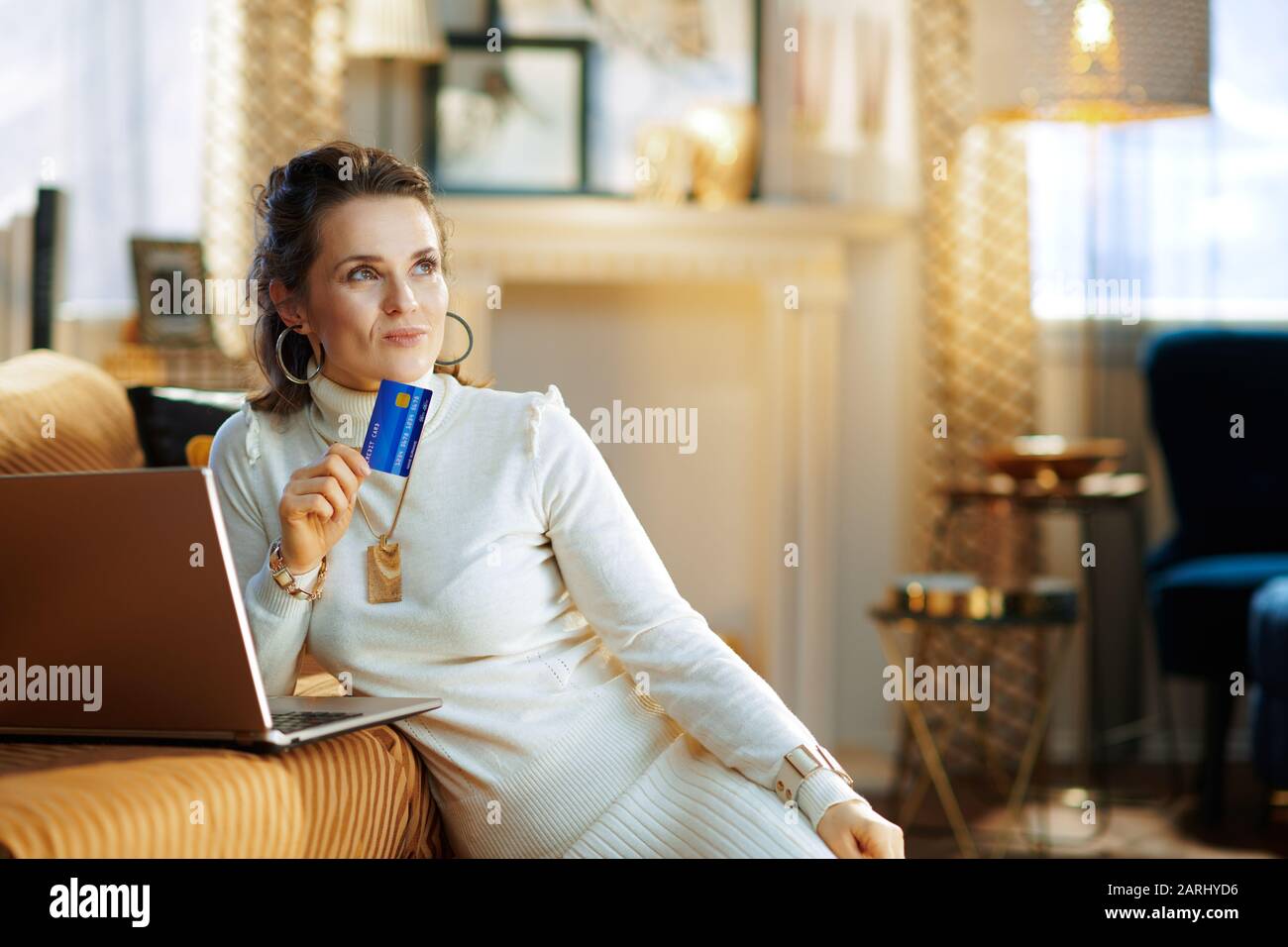 pensive elegant 40 years old housewife in white sweater and skirt with credit card making online purchases on e-commerce website in the modern house i Stock Photo