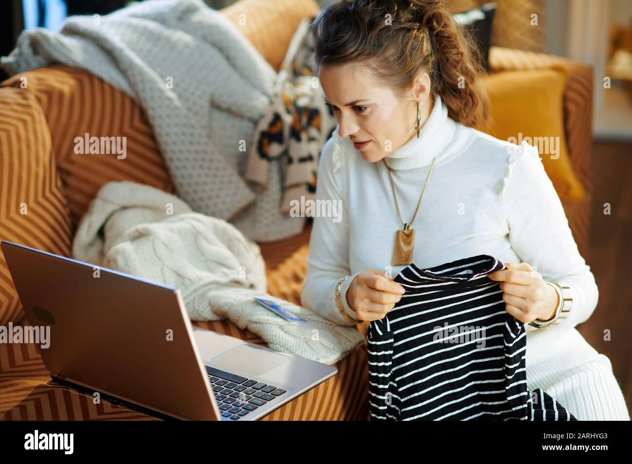 young woman in white sweater and skirt with credit card sitting near couch among old sweaters while buying new sweater shop online on a laptop at mode Stock Photo