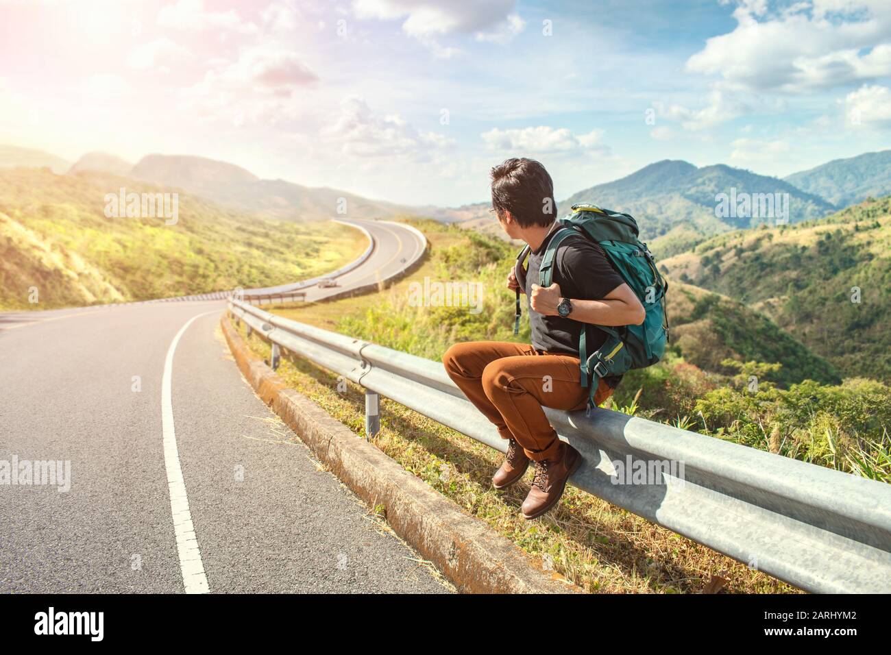 Young man sitting on the roadside. Travel and Holiday concepts. Backpacker on road. Travel man hitchhiking Stock Photo
