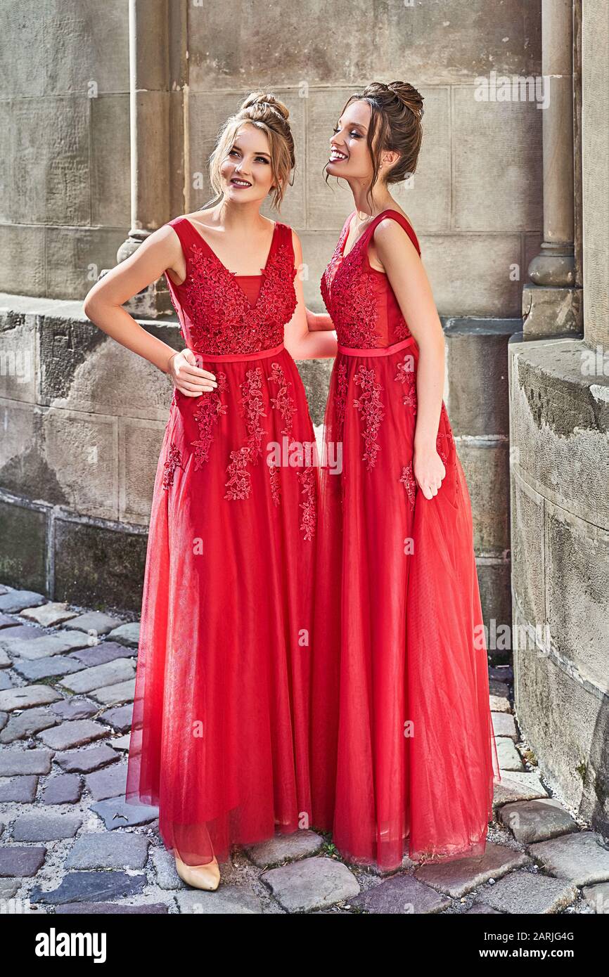 Two beautiful bridesmaids girls blonde and brunette ladies wearing elegant full length red chiffon bridesmaid dress with lace and holding flower Stock Photo