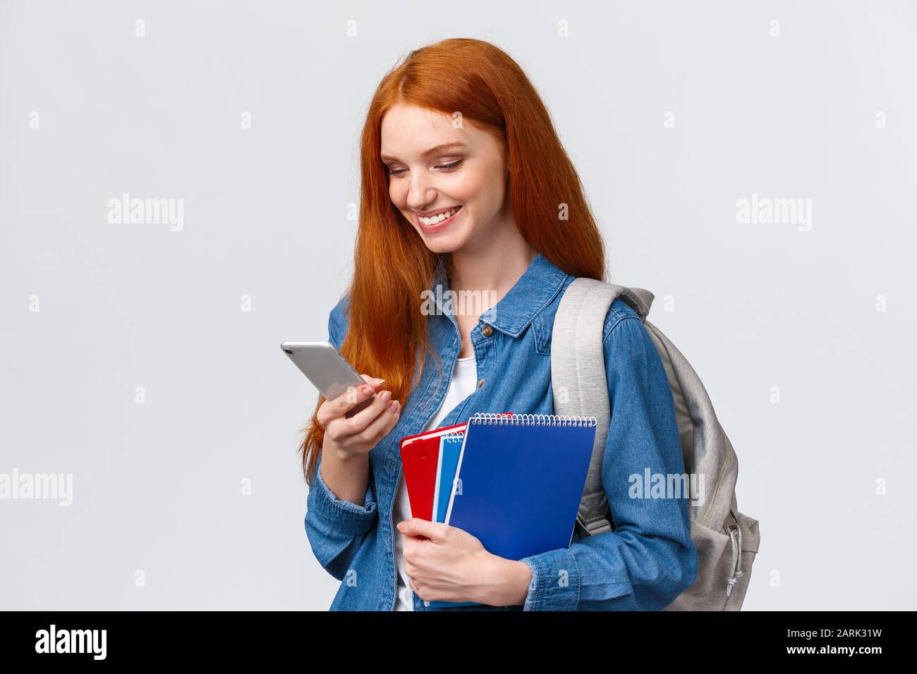 Lovely good-looking female with red hair, heading to college, holding backpack, notebooks smiling as answer friend message, chatting glancing mobile Stock Photo