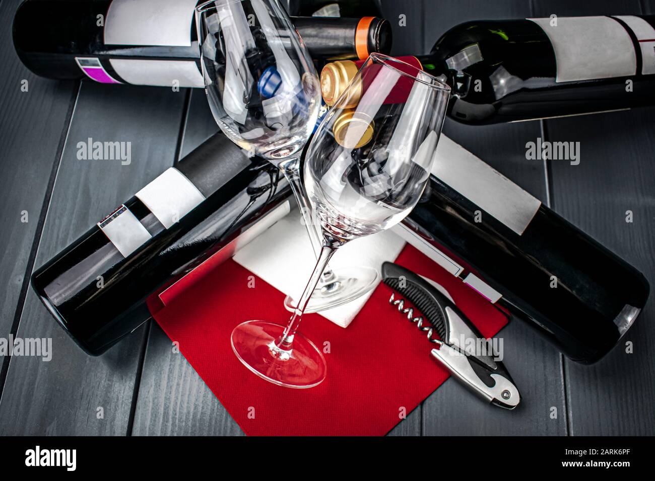 Bright wine glasses lying on bed of bottles of red wine on red tablecloth, corkscrew, napkin and gray wooden background Stock Photo