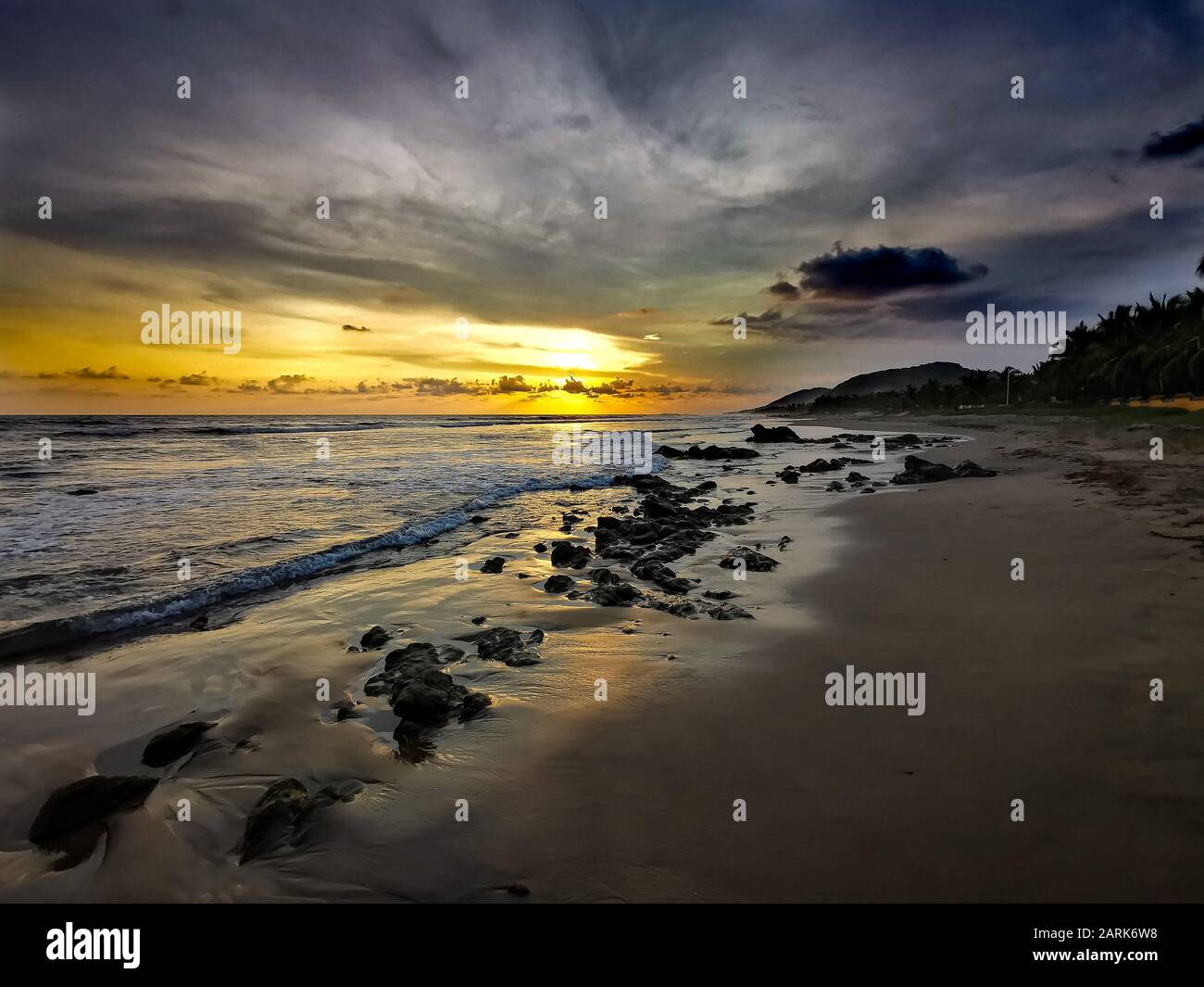 The sun contrasting with the sky, clouds, water, rocks and sand on the Stock Photo