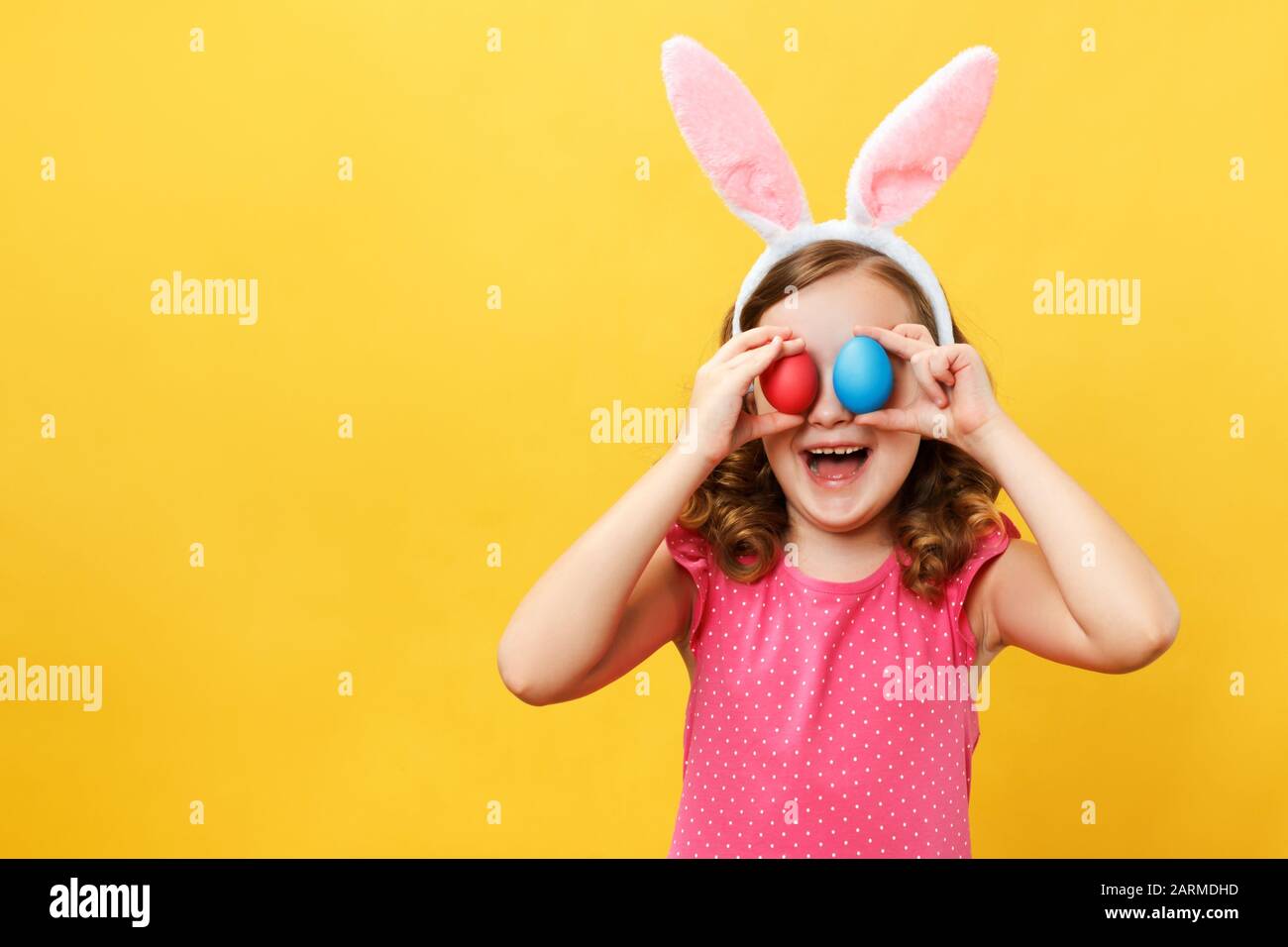Cheerful happy child in the ears of a bunny holds two Easter eggs in his eyes. Portrait of a little girl on a yellow background in the studio. Stock Photo