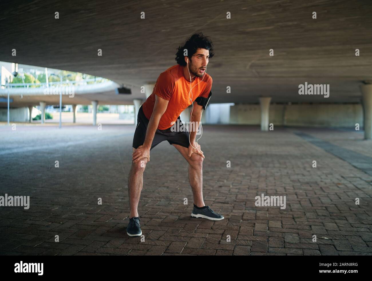 Young male athlete listening to music on earphones feels tired after running and relaxing under the city bridge Stock Photo