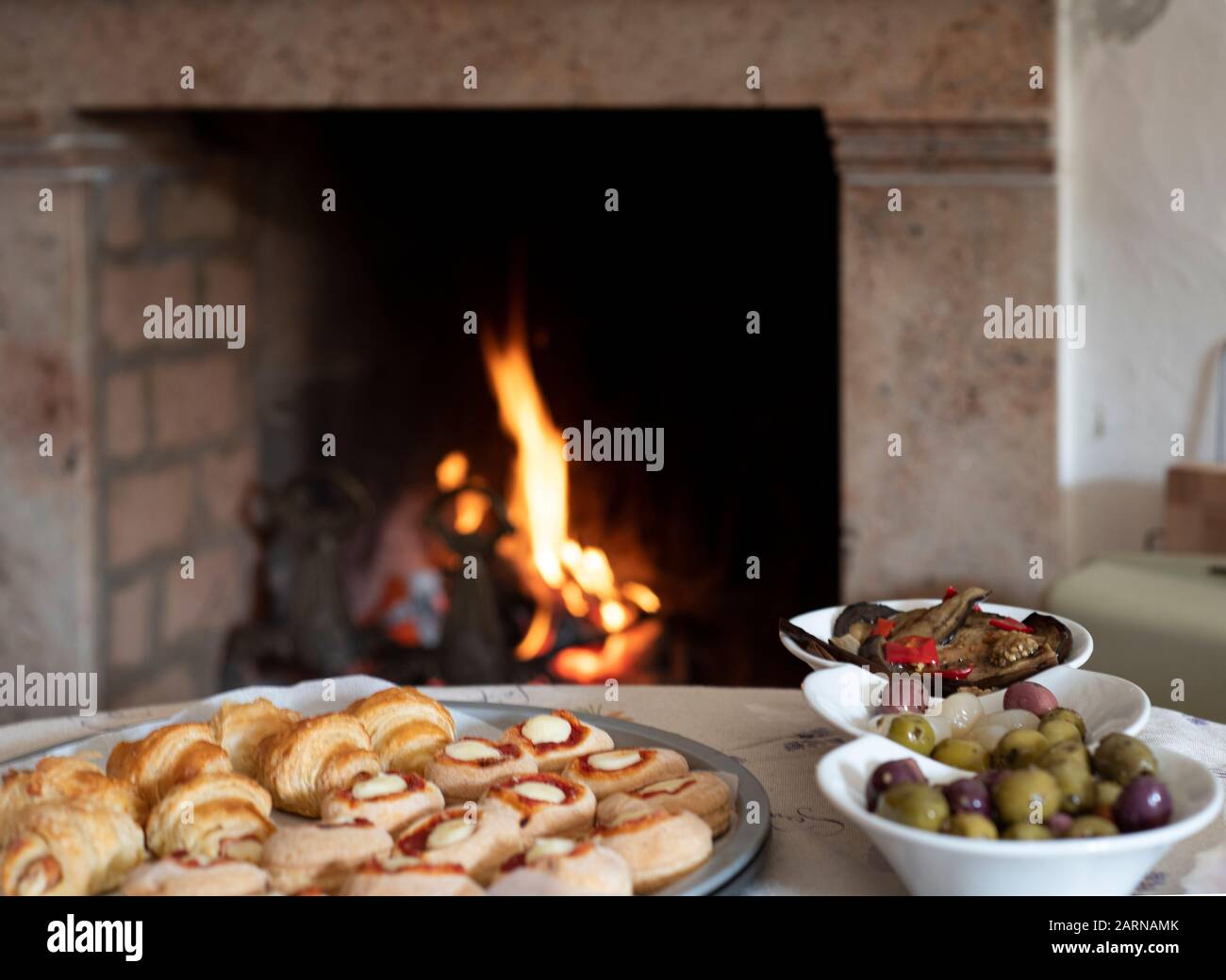Plates of food and olives near a burning fire in a brick hearth indoors in close up with focus to the food Stock Photo