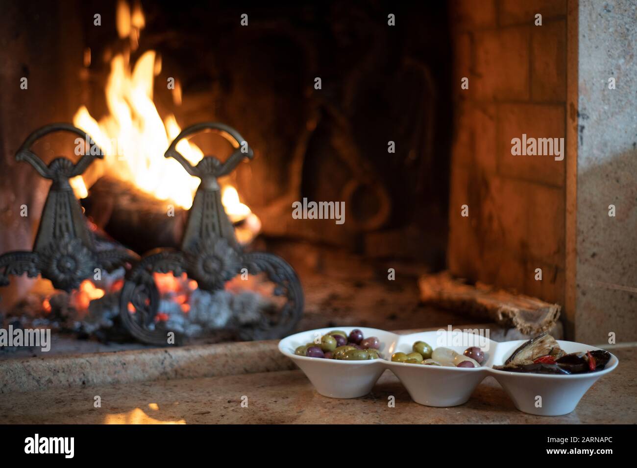 Bowls of Italian olives alongside a burning fire in a brick fireplace on a cold winter day in close up Stock Photo