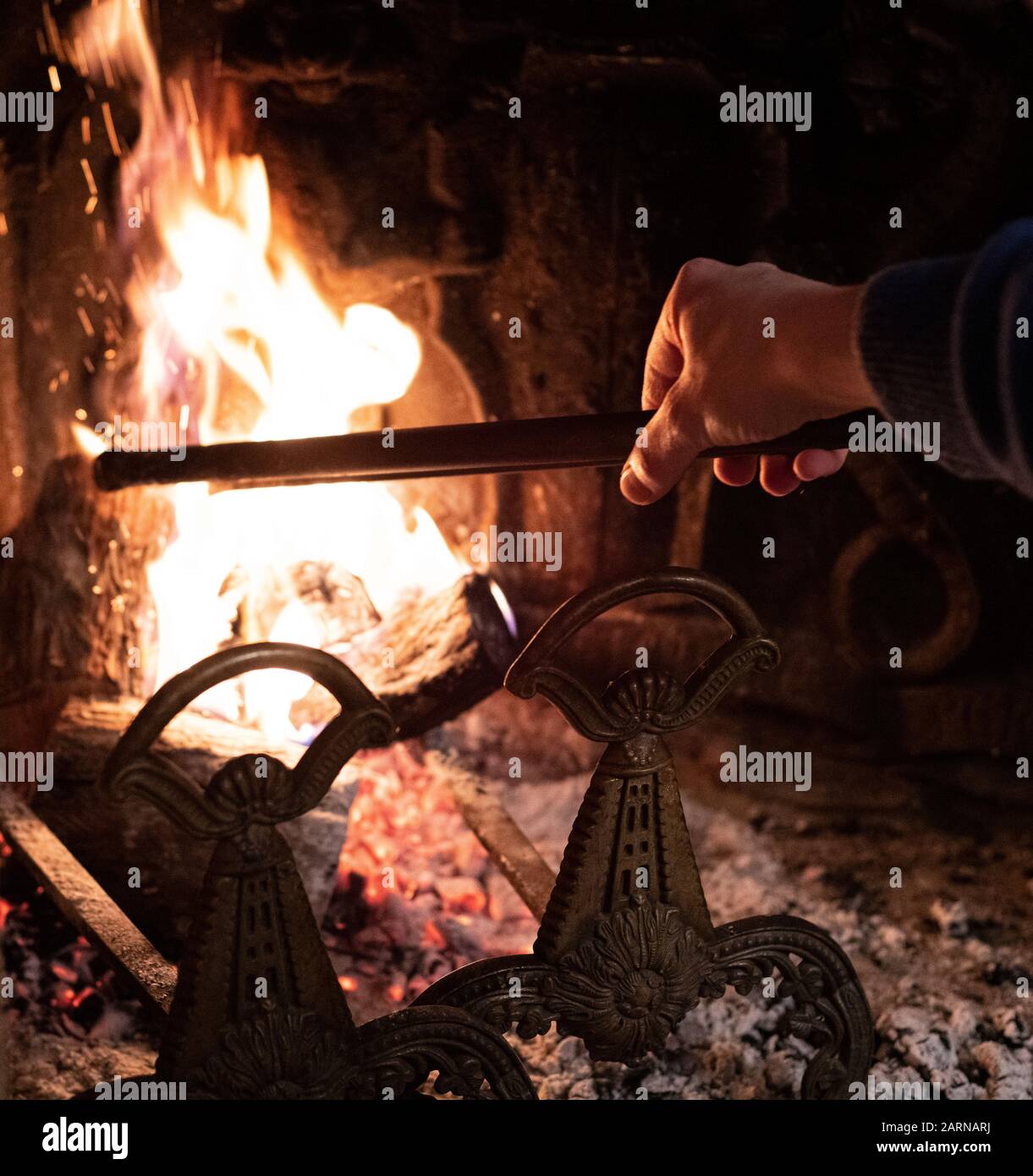 Person using a fire iron to stoke the flames on a fire burning in an indoor brick fireplace behind andirons in a close up view on the flames Stock Photo