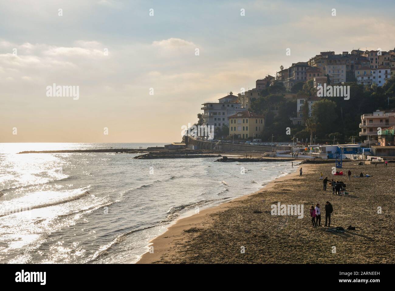 Sandy beach of Porto Maurizio with people enjoying the sun in a warm winter day and the Parasio old borough on the cape, Imperia, Liguria, Italy Stock Photo