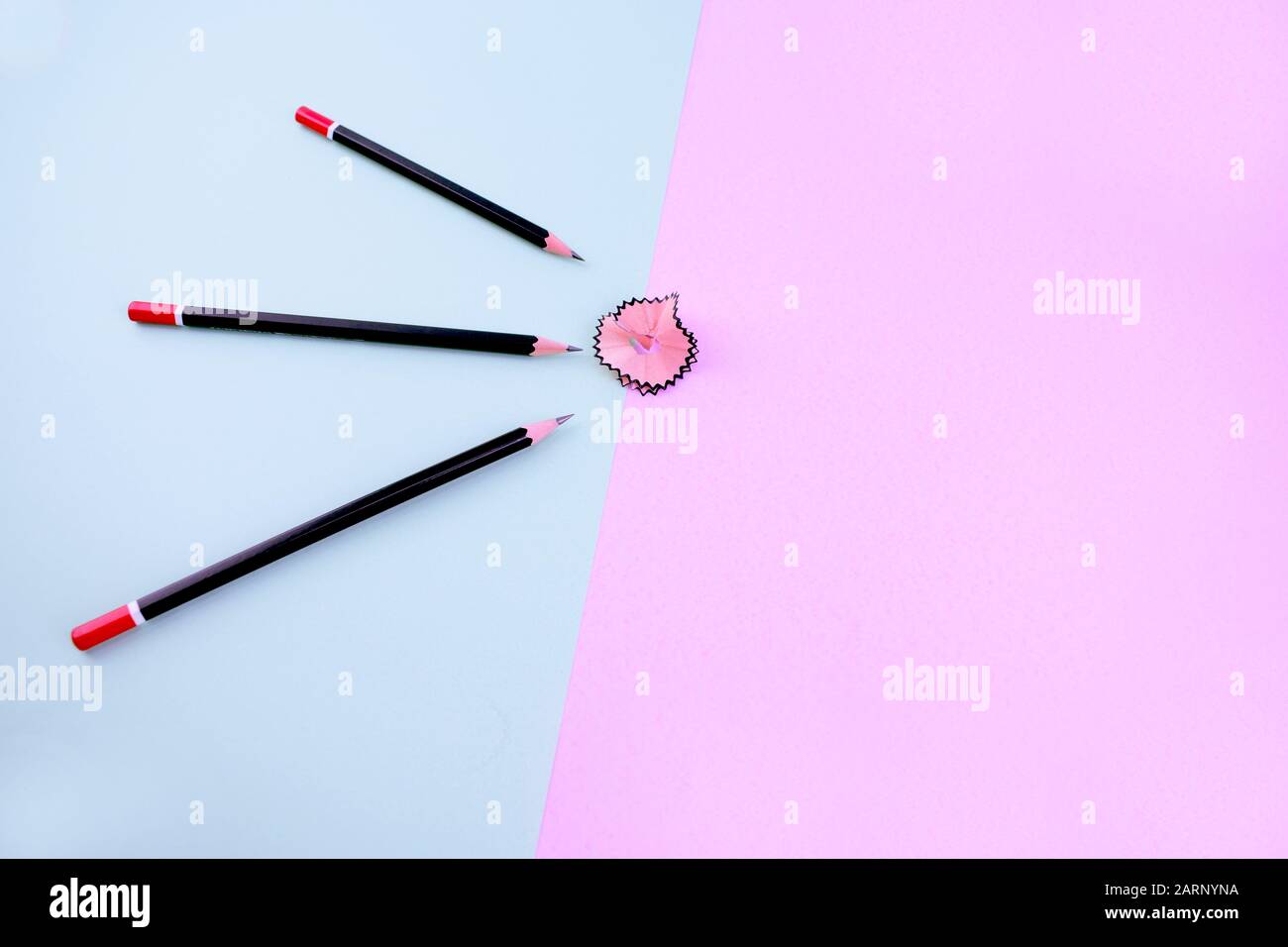 three dark black sharpened pencils over a light blue background are pointing at a pink pencil shaving which on a pink paper background Stock Photo