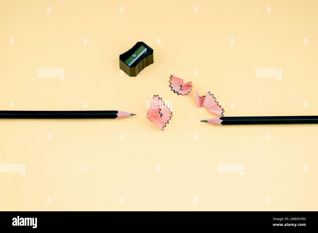 couple of dark black sharpened wooden pencil lying beside a sharpener and some pencil shavings over a light orange paper background Stock Photo