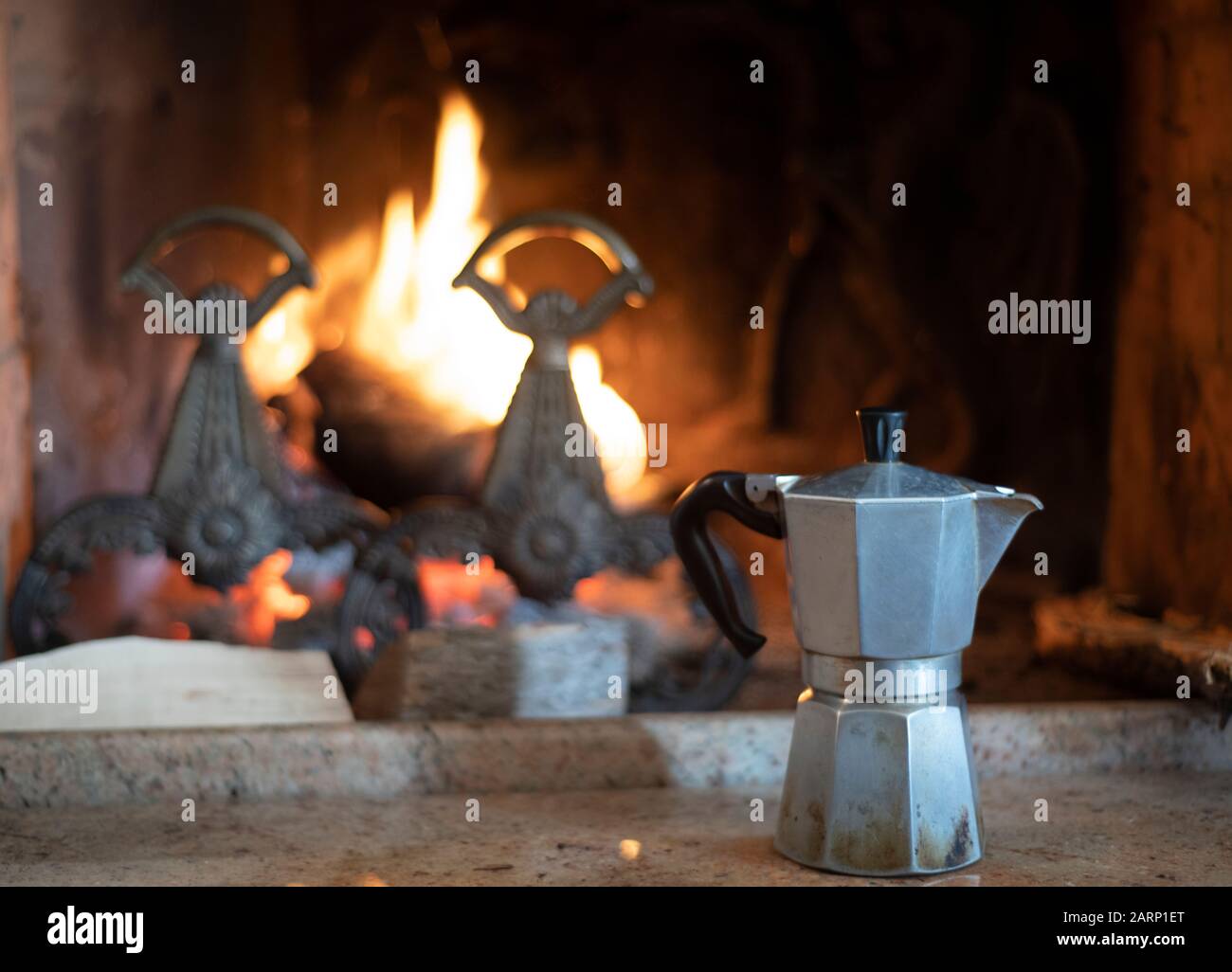 Italian coffee pot in front of a burning fire with andirons in a brick indoor chimney in a concept of relaxation Stock Photo